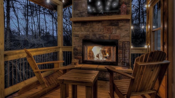 A cozy cabin interior with a stone fireplace, wooden furniture, and a view of the forest. - Wolfs Den II - Shuffleboard | Foosball | Fire Pit | Outdoor Wood Burning Fireplace (Blue Ridge)