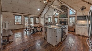 A spacious kitchen with a central island, wooden flooring, and a stone fireplace. - Wolfs Den II - Shuffleboard | Foosball | Fire Pit | Outdoor Wood Burning Fireplace (Blue Ridge)