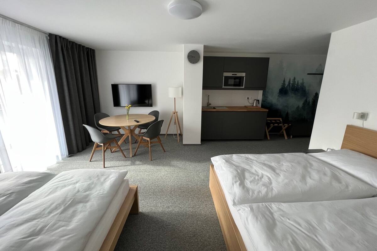 Ein Hotelzimmer mit zwei Betten, einem Esstisch mit Stühlen, einem Fernseher, einer kleinen Küchenzeile und einem Waldgemälde.