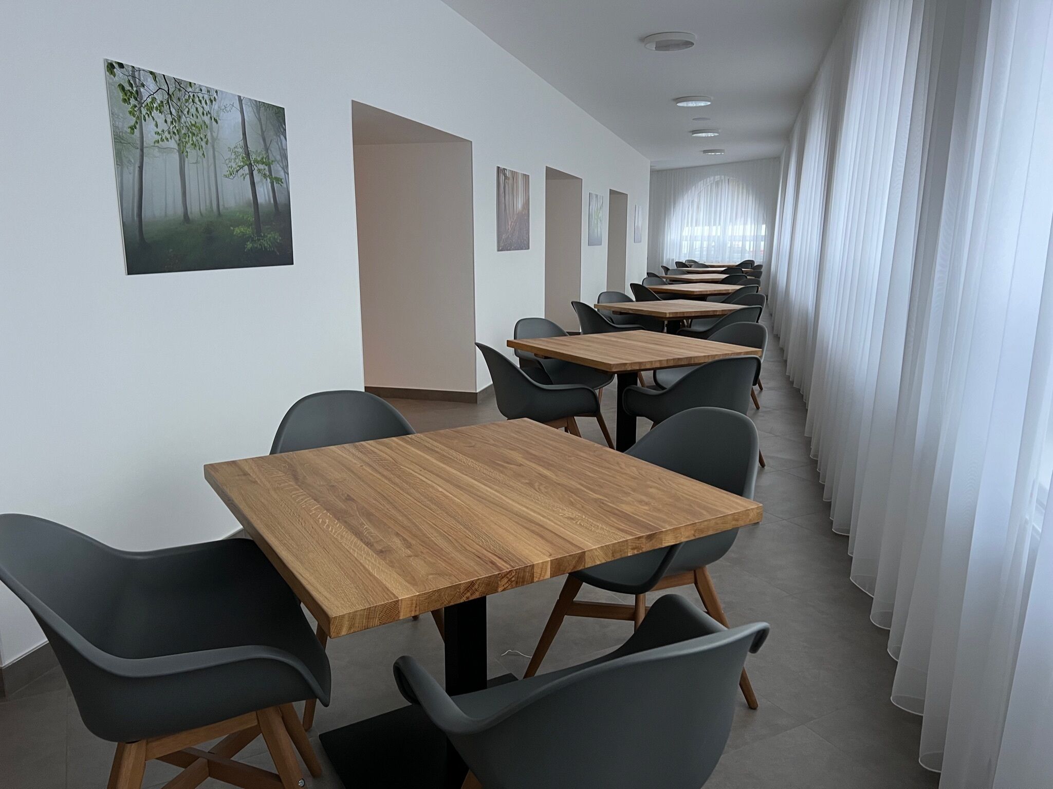 A long corridor with multiple wooden tables and black chairs, white walls, and a large window with white curtains.