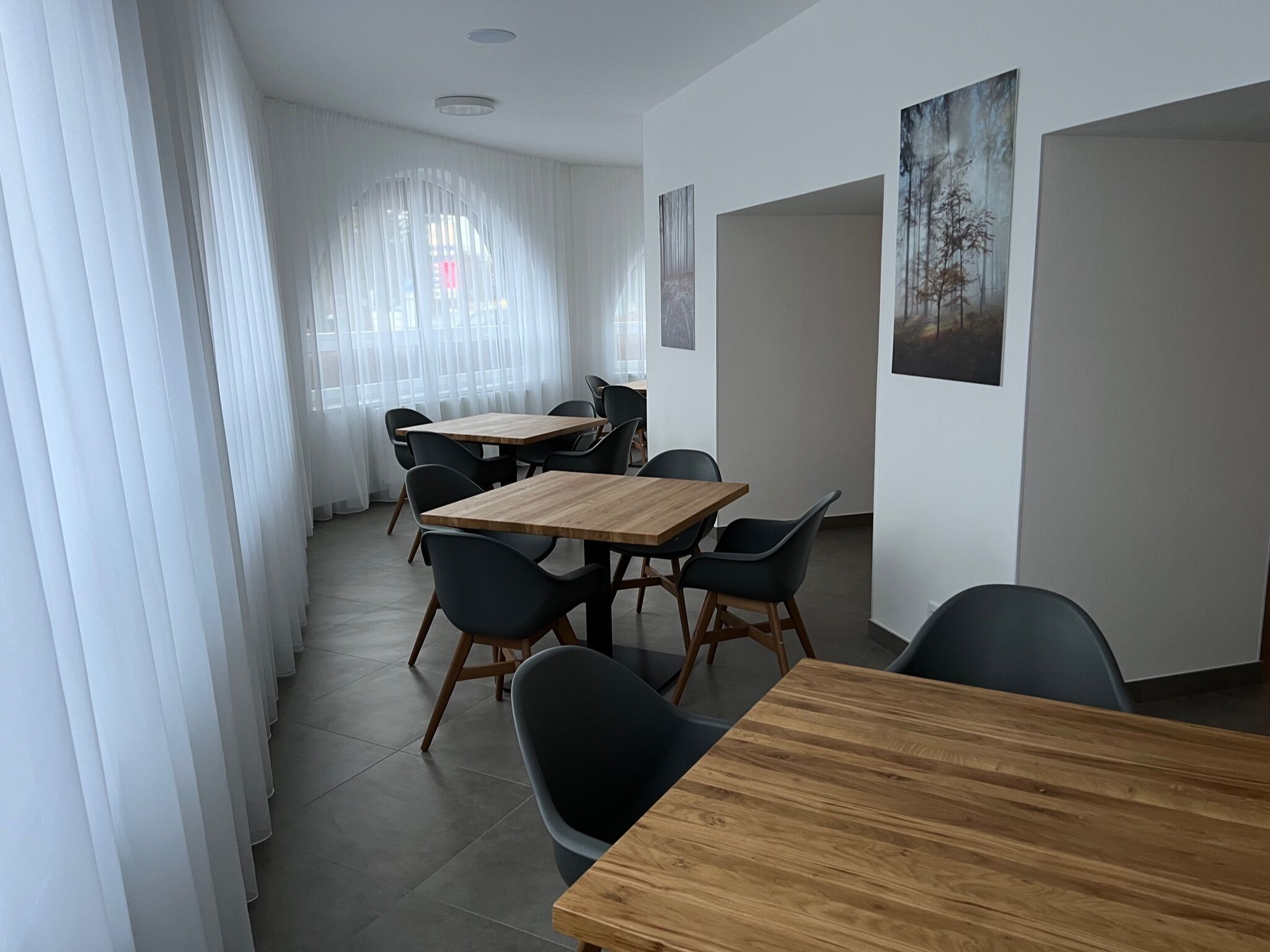 Una sala de reuniones moderna con mesas de madera y sillas negras, ventanas amplias con cortinas ligeras y obras de arte enmarcadas en las paredes.