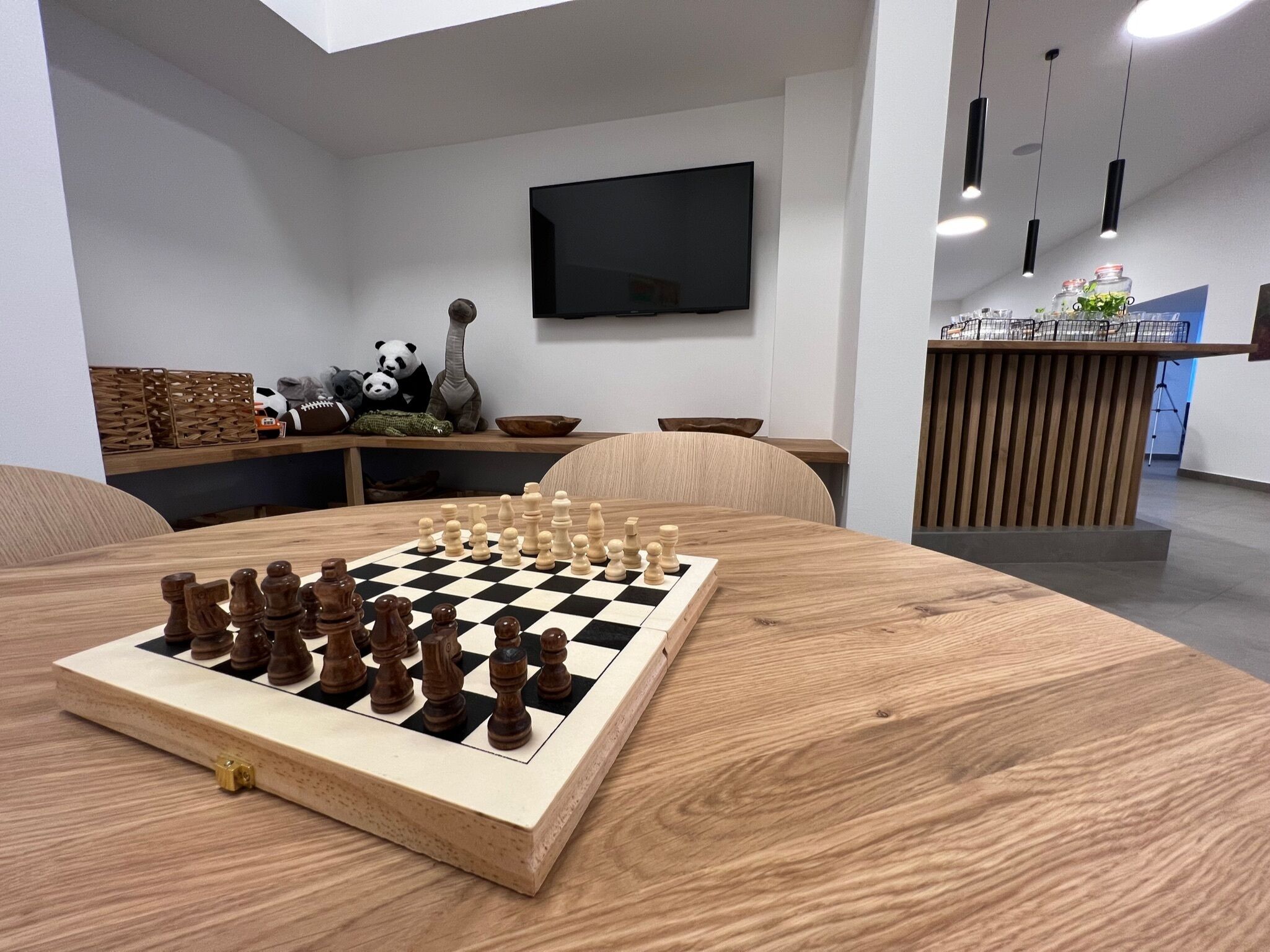 A wooden chess table in a modern living room with a TV, wooden furniture, and a kitchen area in the background.