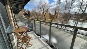 Un balcón con mesa y sillas de madera, barandilla metálica y vista a un lago y árboles.
