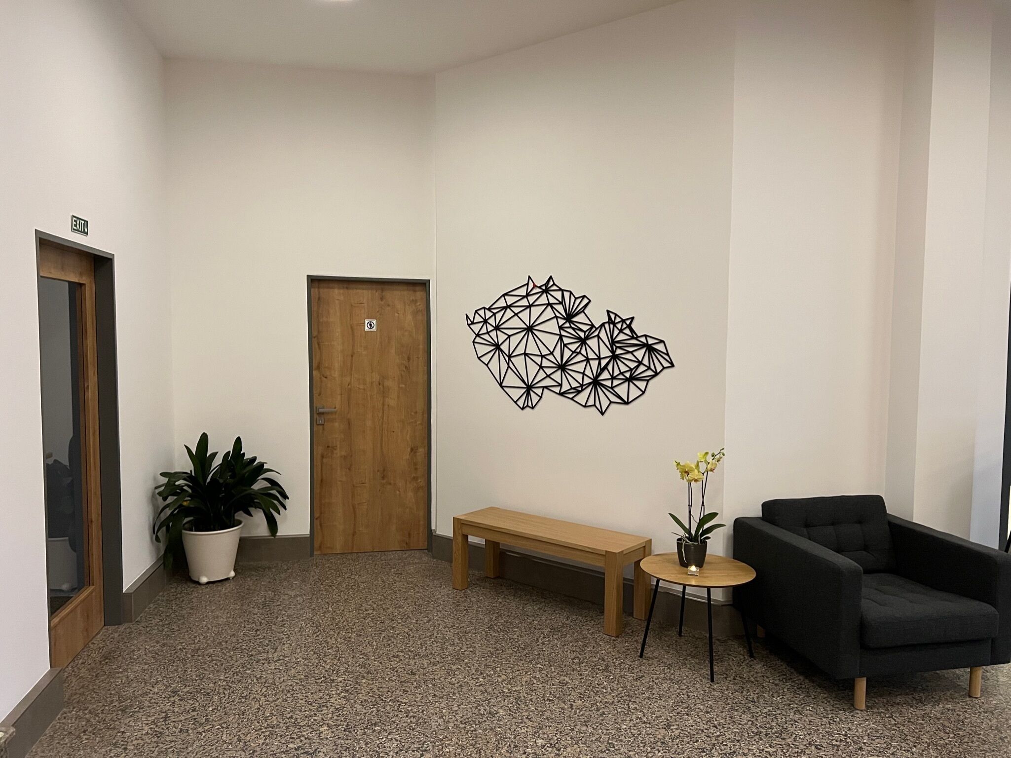 Uma área de espera moderna com uma porta de madeira, um banco, uma poltrona preta, uma pequena mesa redonda com um vaso de planta e um ornamento geométrico na parede.
