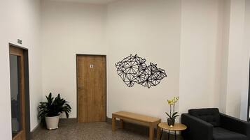 Uma área de espera moderna com uma porta de madeira, um banco, uma poltrona preta, uma pequena mesa redonda com um vaso de planta e um ornamento geométrico na parede.