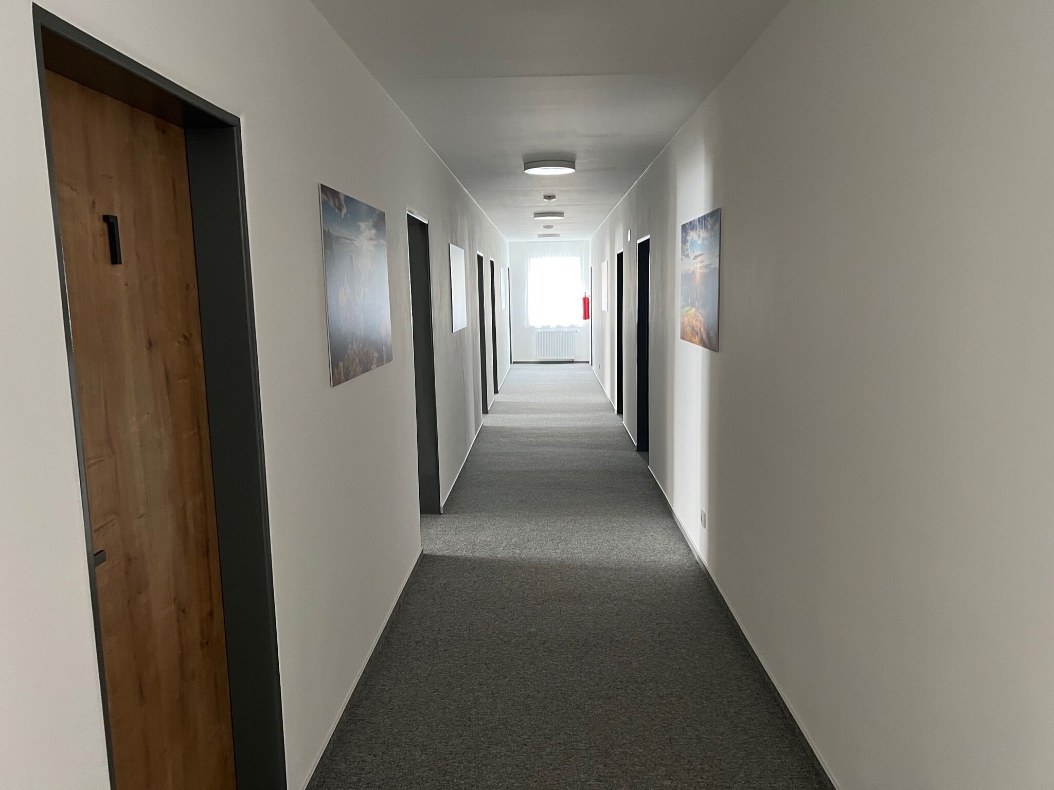 Um corredor comprido e estreito, com pisos cobertos de carpete e paredes brancas. Há portas em ambos os lados, algumas com painéis de madeira.
