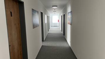 Um corredor comprido e estreito, com pisos cobertos de carpete e paredes brancas. Há portas em ambos os lados, algumas com painéis de madeira.