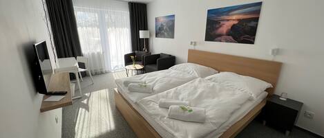 A hotel room with a large bed, a desk with a laptop, a chair, a small table with a vase, and two paintings on the wall.