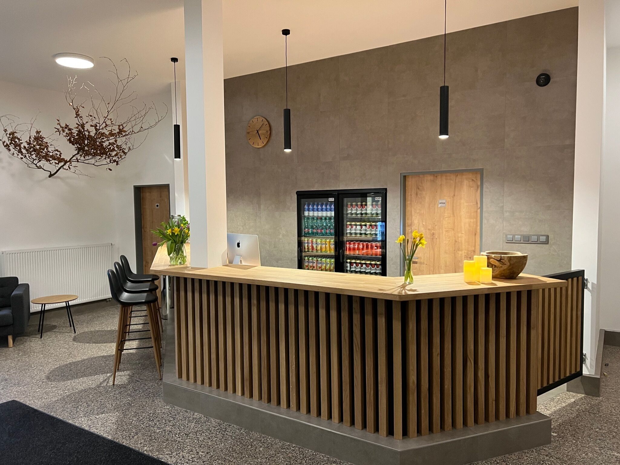 Un espace d’accueil comprenant un comptoir de réception en bois, deux lampes suspendues, une horloge, un réfrigérateur rempli de boissons et un vase avec des fleurs.