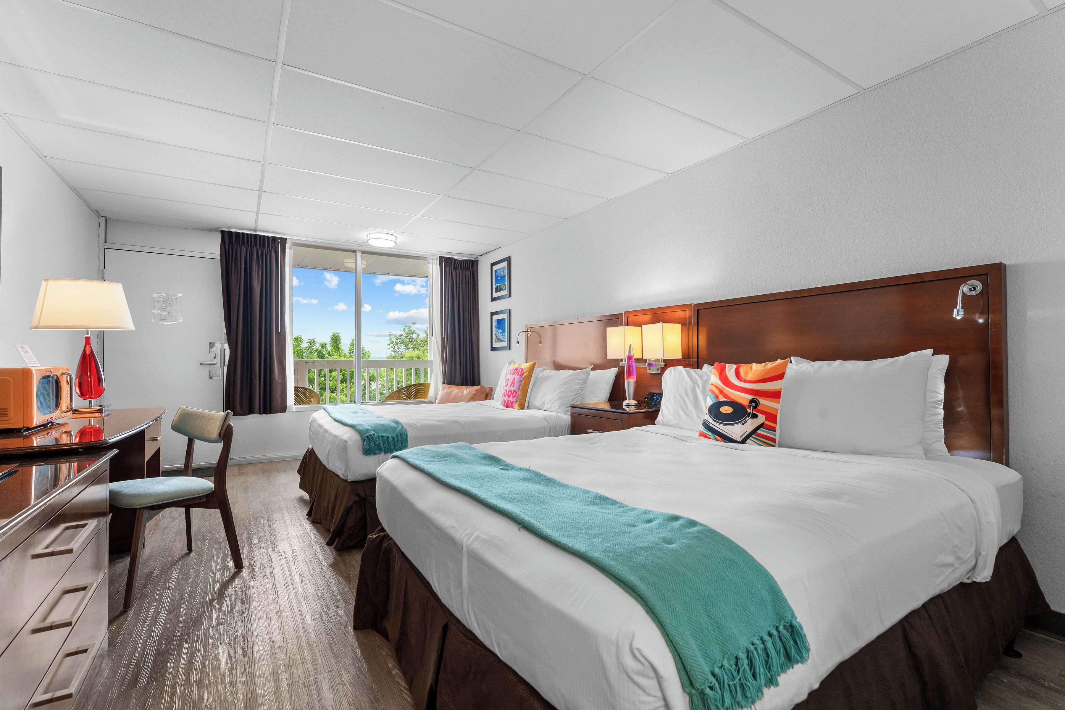A hotel room with two beds, a desk with a lamp, a chair, and a view of the outdoors.