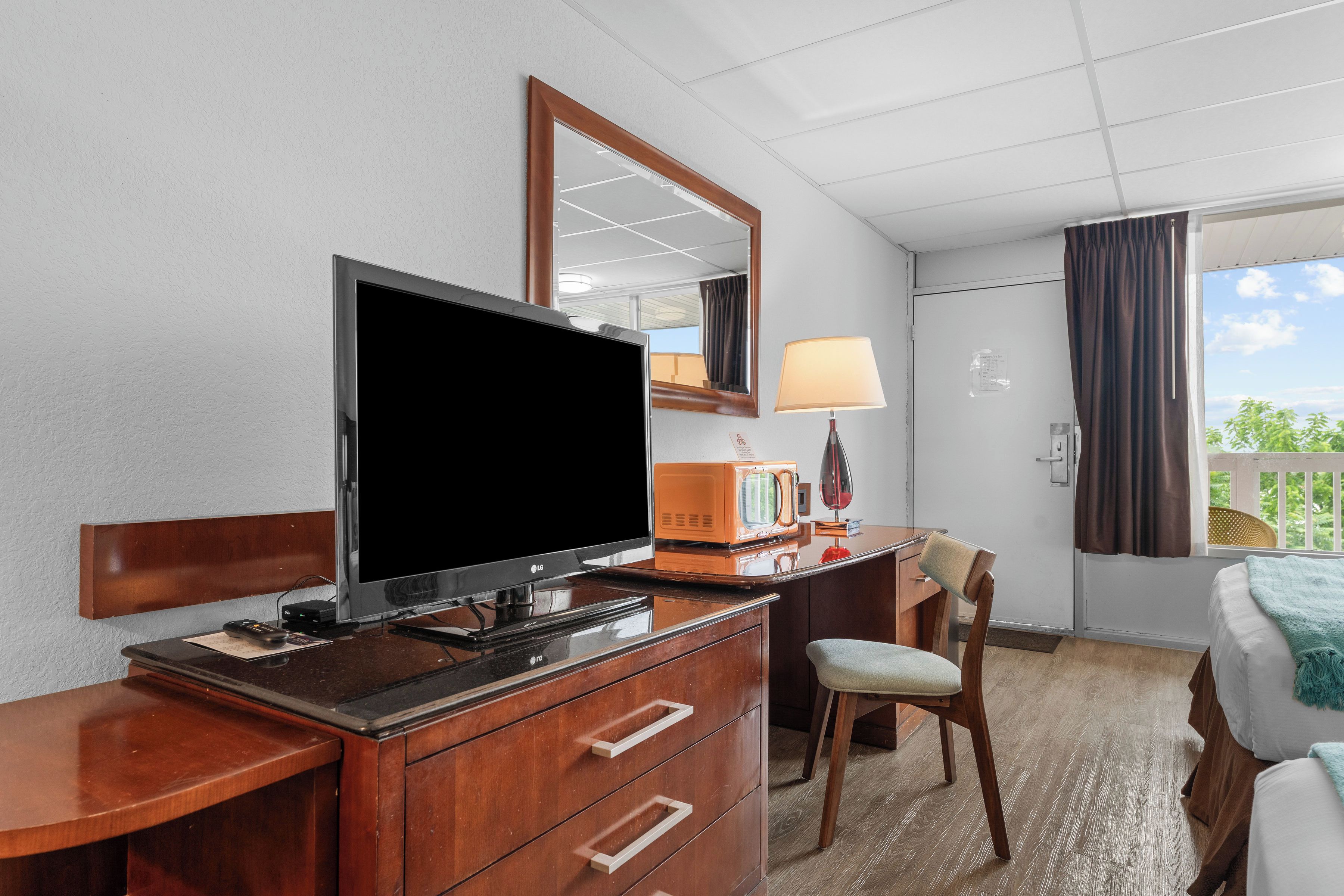 Habitación de hotel con televisor de pantalla plana, cómoda de madera, lámpara, una silla y una cama con vistas al exterior.