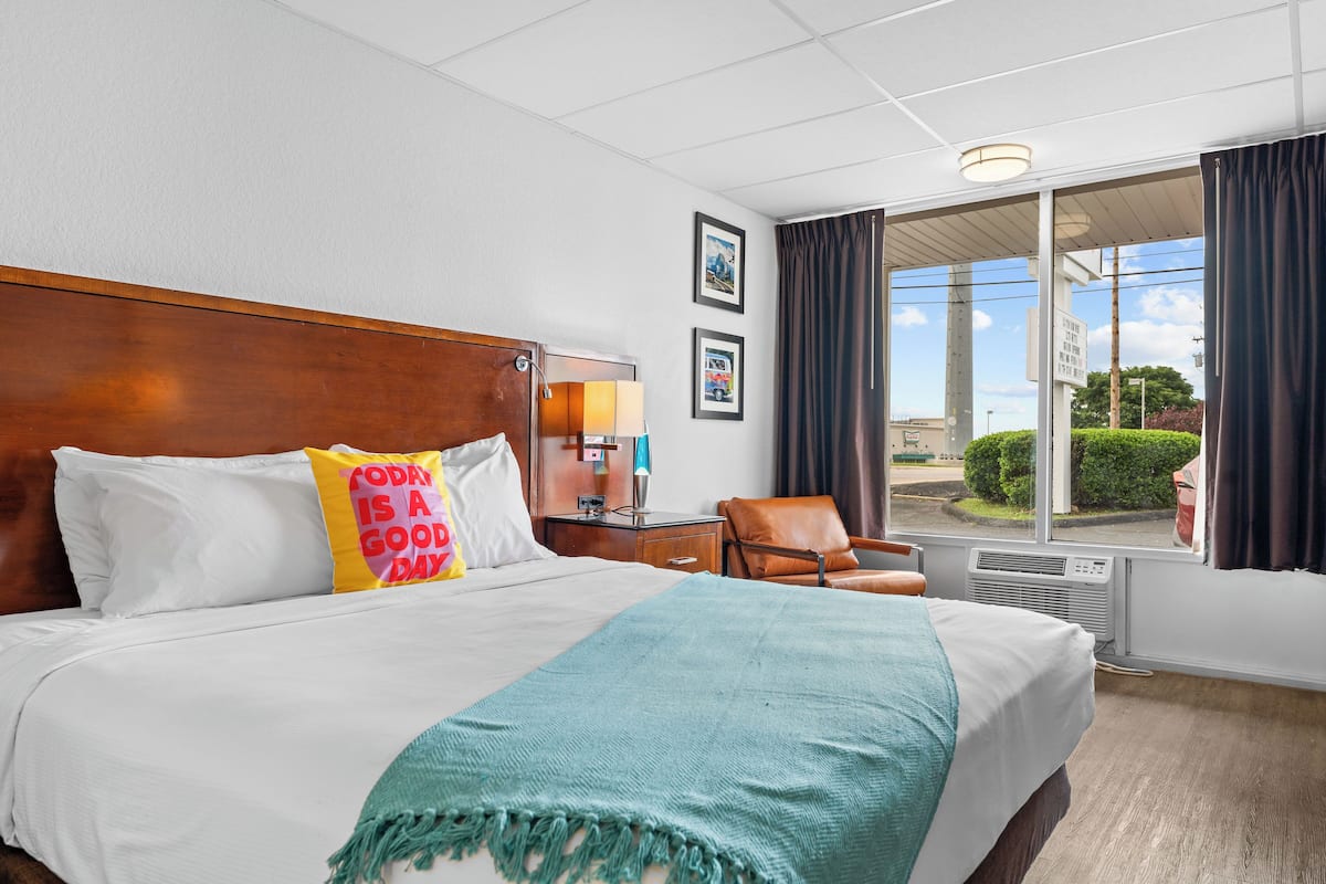 A hotel room with a large bed, a nightstand with a lamp, a chair, and a view of the outdoors through a window.