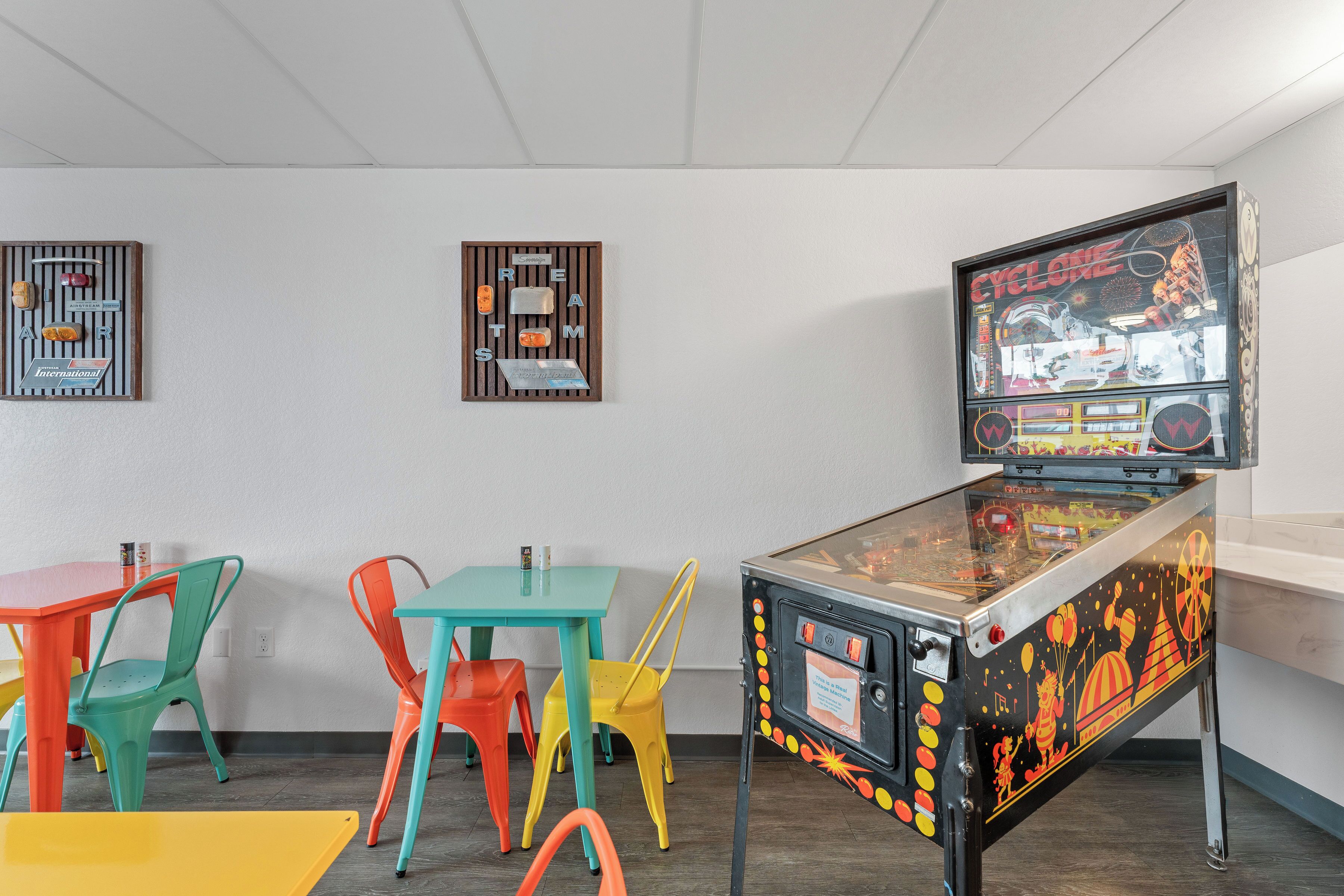 Uma máquina de pinball, cadeiras coloridas e uma mesa numa sala com paredes brancas.