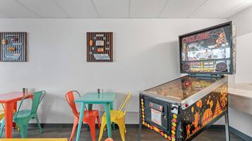 Uma máquina de pinball, cadeiras coloridas e uma mesa numa sala com paredes brancas.