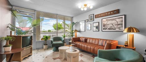 A modern living room with a sofa, armchairs, a coffee table, and a TV. There are plants, a lamp, and artwork on the walls.