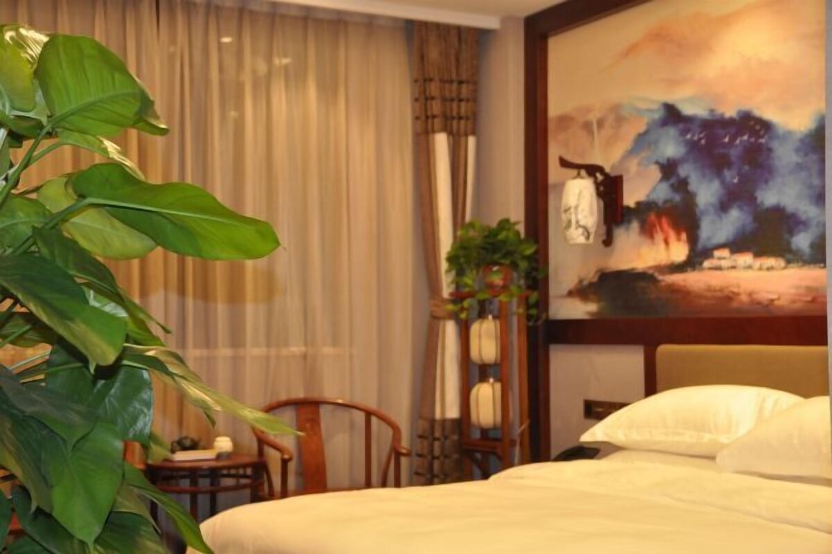 Una habitación de hotel con una cama, una silla, una mesa, una planta en maceta, un cuadro y cortinas.