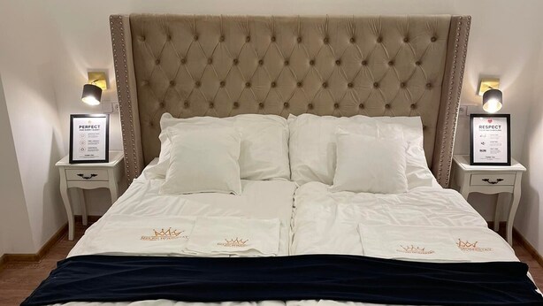 A neatly made bed with a tufted headboard, white bedding, and a dark blue throw blanket. There are two bedside tables with framed signs and lamps. A bench is placed in front of the bed.