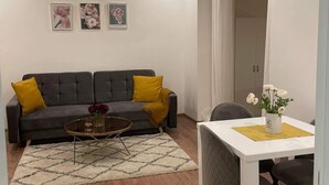 A modern living room with a grey sofa, yellow cushions, a coffee table, and a dining table with chairs. There are three framed floral pictures on the wall.
