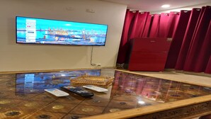 Quarto com televisão de tela plana na parede exibindo uma vista urbana, uma mesa de centro de vidro com controles remotos, uma geladeira vermelha e cortinas vermelhas.