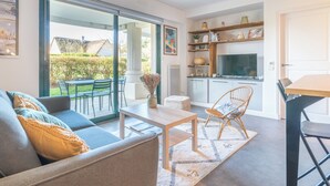 Ein modernes Wohnzimmer mit Sofa, Couchtisch und Stühlen, eine Küche mit Fernseher sowie ein Blick ins Freie.