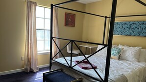 Una cama con dosel, un banco, una mesita y un ventanal con cortinas.