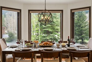 A dining table set with plates, glasses, and a bottle of wine, with a view of trees through large windows.