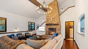 A modern living room with a stone fireplace, a large sofa, a coffee table, and a chandelier. - Spacious East Vail Getaway by Vail Butler (Vail)
