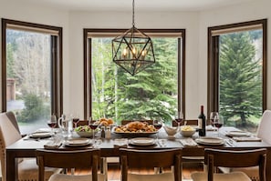A dining table set for a meal with a chandelier, large windows, and a view of trees.