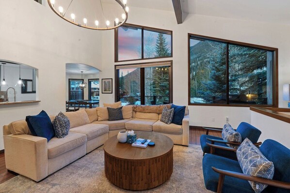 A modern living room with a sectional sofa, a round coffee table, and a large window offering a view of trees.