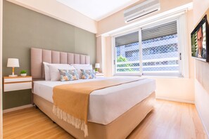 Quarto com uma cama grande, janela com persianas, TV fixada na parede e uma pequena mesa de cabeceira com abajur.
