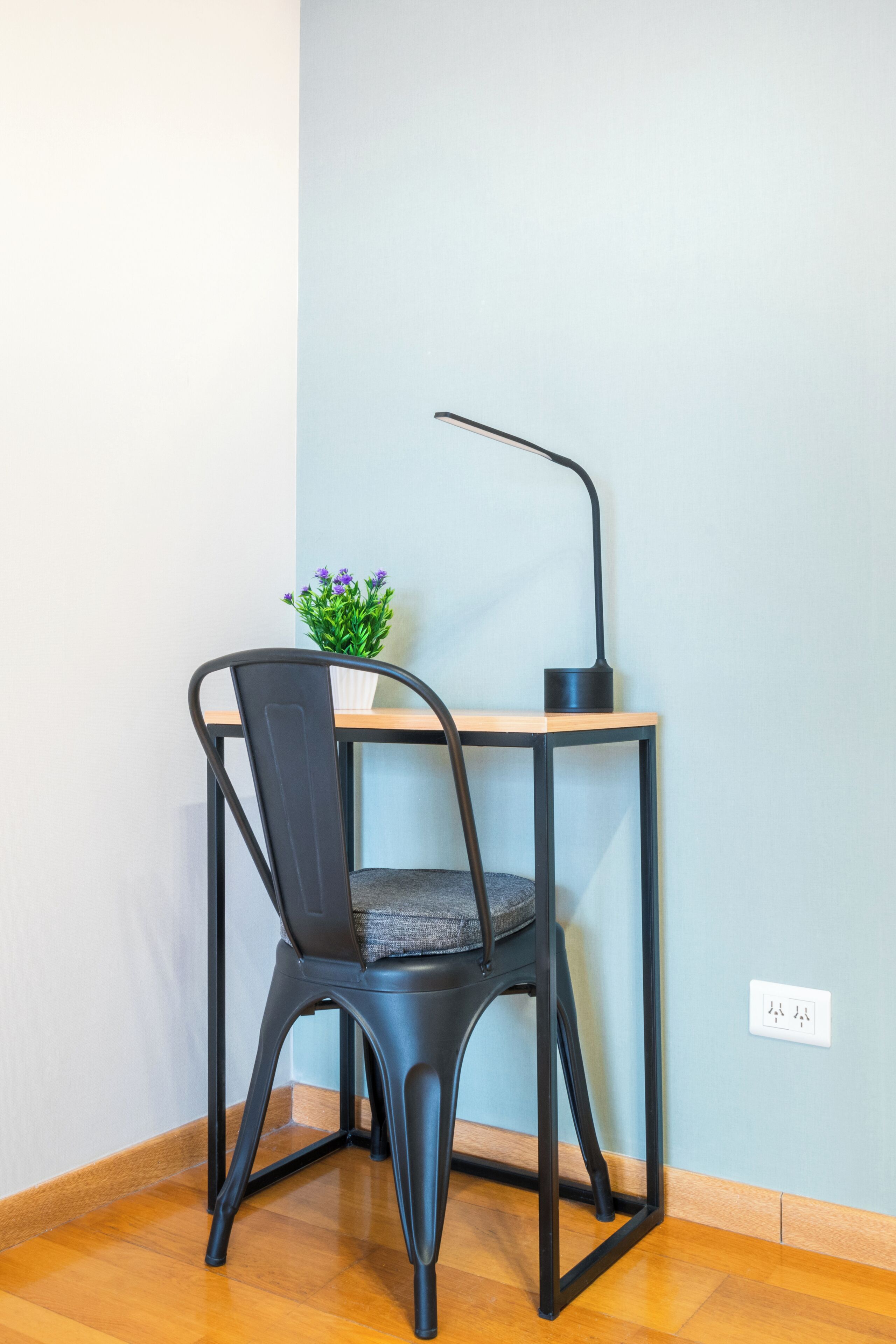 A corner room with a wooden floor, a black metal chair, a small table with a potted plant, and a black desk lamp.