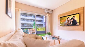 Una sala de estar con sofá, una mesa de centro con libros, un televisor en la pared y un balcón con vistas a edificios.
