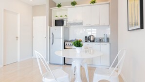Una cocina moderna con armarios blancos, un refrigerador de acero inoxidable, un microondas y una estufa. Hay una mesa de comedor blanca con dos sillas y una planta en maceta encima.