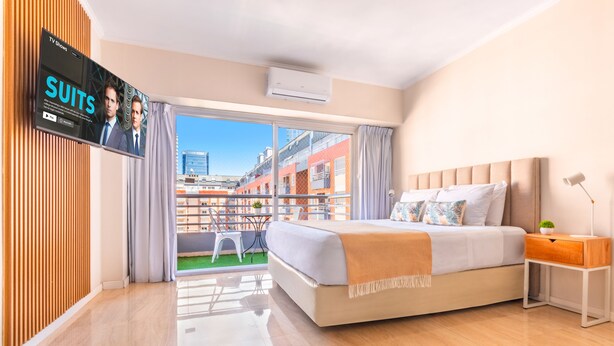 Habitación de hotel moderna con una cama grande, un televisor montado en la pared y un balcón con vistas a la ciudad.