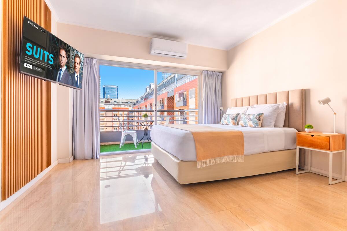Habitación de hotel moderna con una cama grande, un televisor montado en la pared y un balcón con vistas a la ciudad.