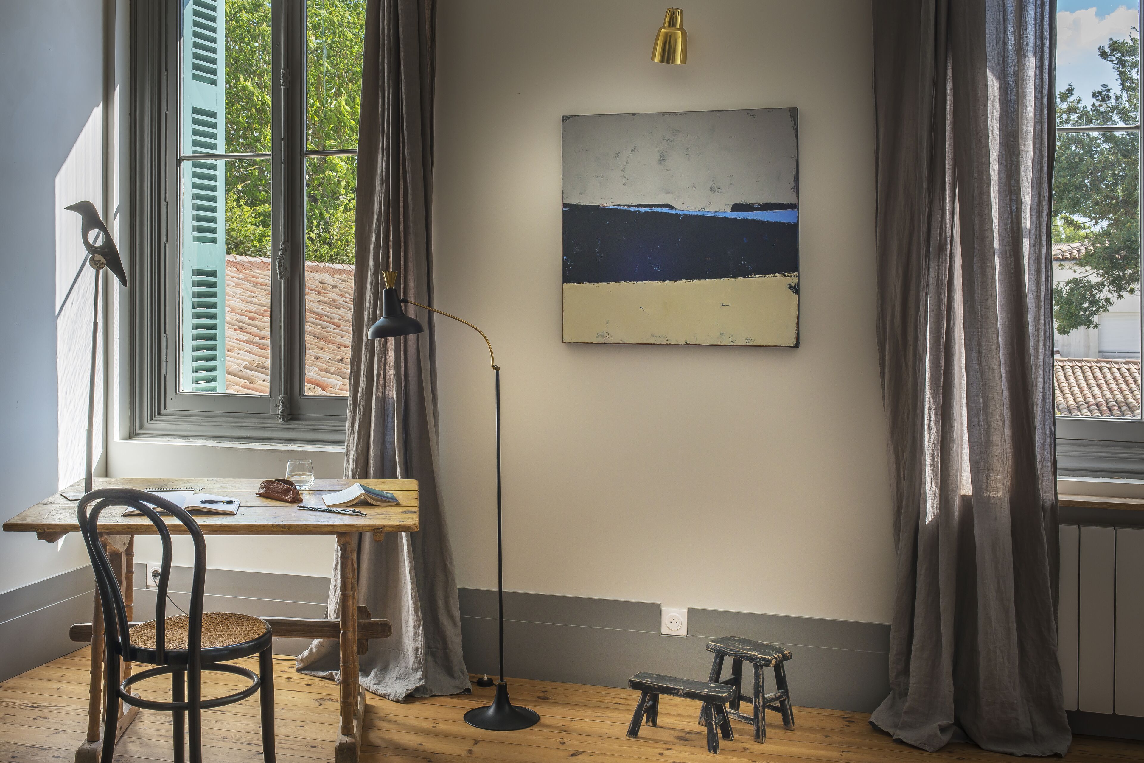 Una habitación con un escritorio de madera, una silla, un taburete pequeño, una lámpara de pie y un cuadro en la pared.