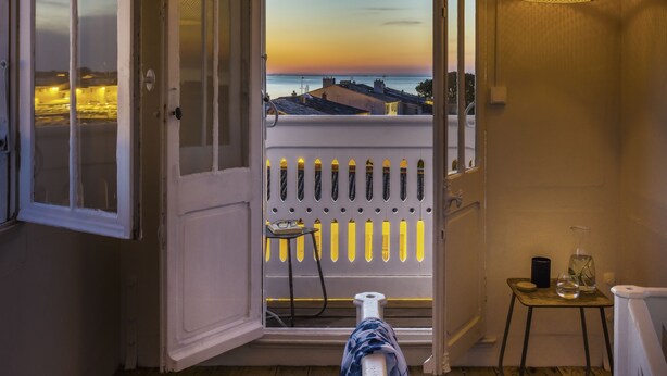 A room with a wooden ceiling, a balcony with a railing, a hanging light fixture, and a view of the sunset. - LE LANTERNON (Saint-Martin-de-Re)