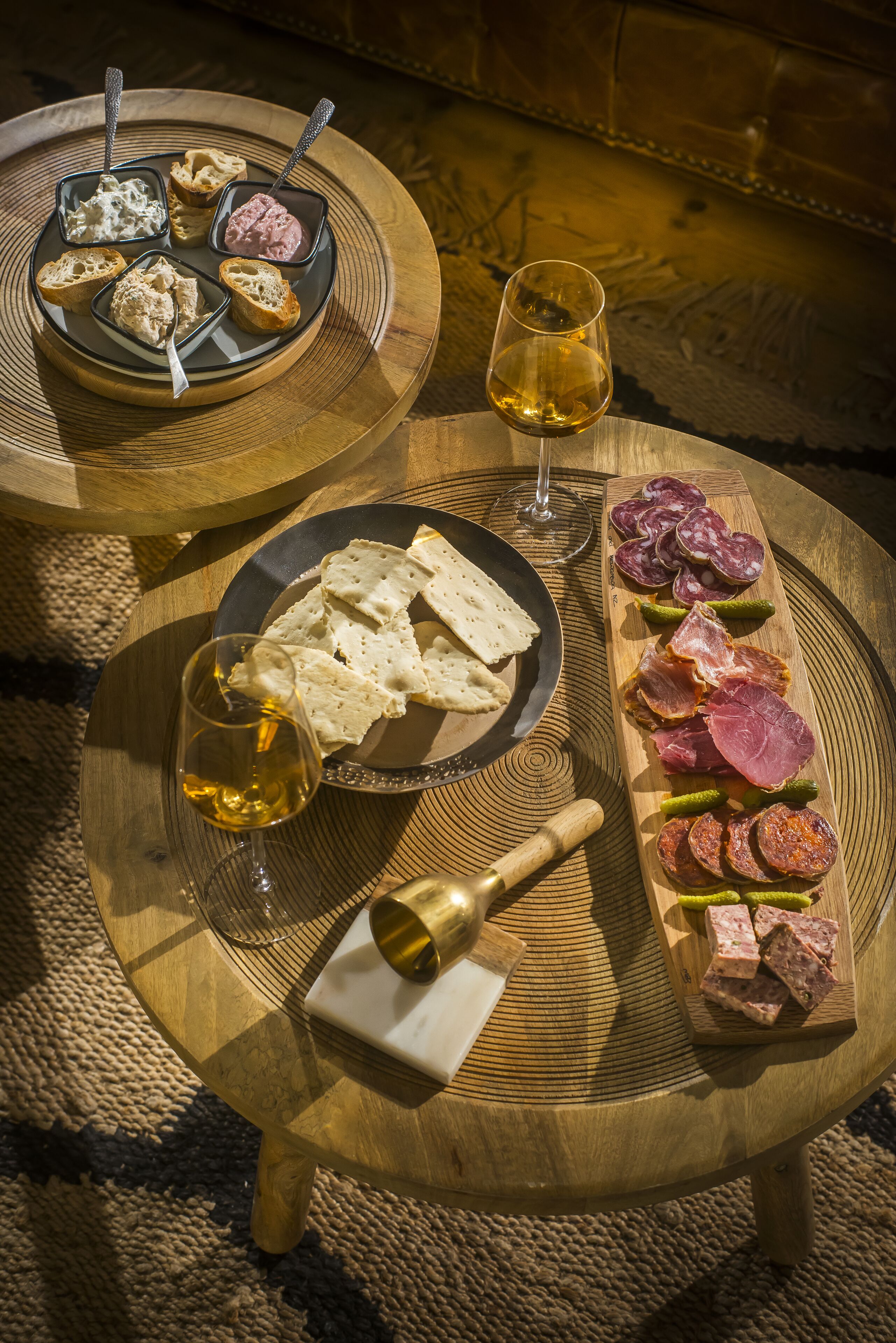 Dos mesas redondas de madera con comida y bebidas. Una mesa tiene pan y queso, y la otra, carnes y encurtidos.