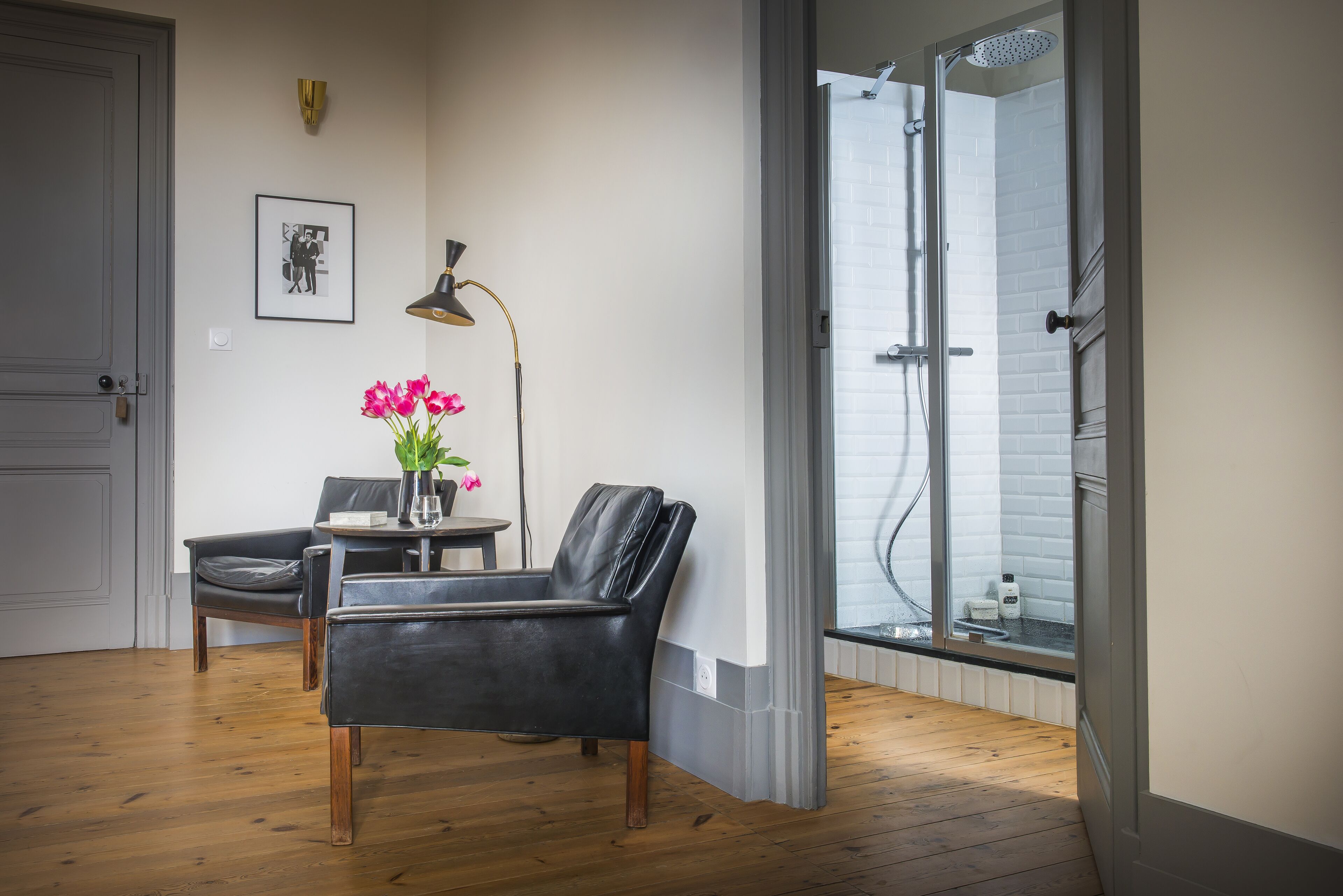 A room with a black leather chair, a small table with a vase of pink flowers, a black floor lamp, and a window with a shower visible through the glass.