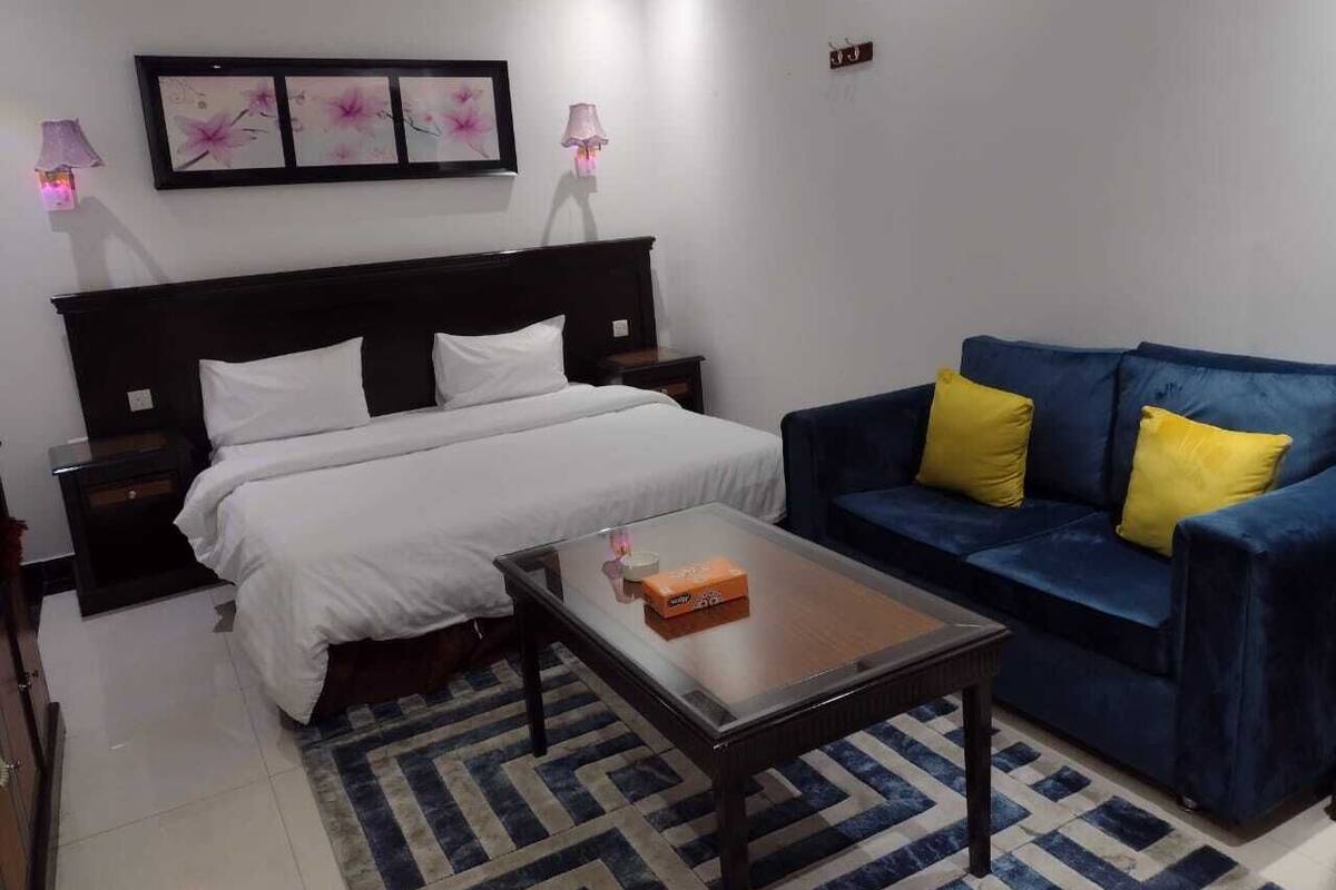 A hotel room with a bed, a blue sofa with yellow pillows, a coffee table, and a framed floral artwork on the wall.