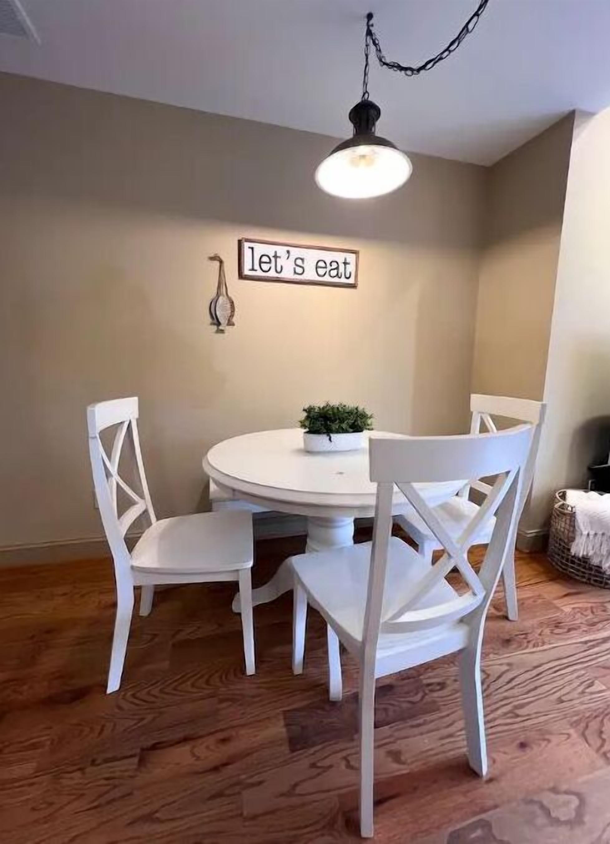A dining area with a round table, four white chairs, a hanging light fixture, and a wall sign that reads "let's eat."