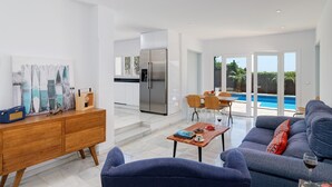 Ett modernt vardagsrum med en blå soffa, ett träbord och en matplats med stolar och ett bord. Det finns ett kök med ett kylskåp och ett matbord. En pool syns genom glasdörrarna.