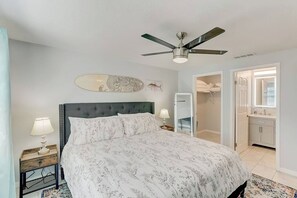 Une chambre à coucher comprenant un lit, un ventilateur de plafond, une table de chevet avec une lampe, un tableau décoratif au mur et une armoire avec des vêtements suspendus.