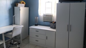 Una habitación con escritorio blanco, una silla, armario blanco, televisor y ventana con cortinas.