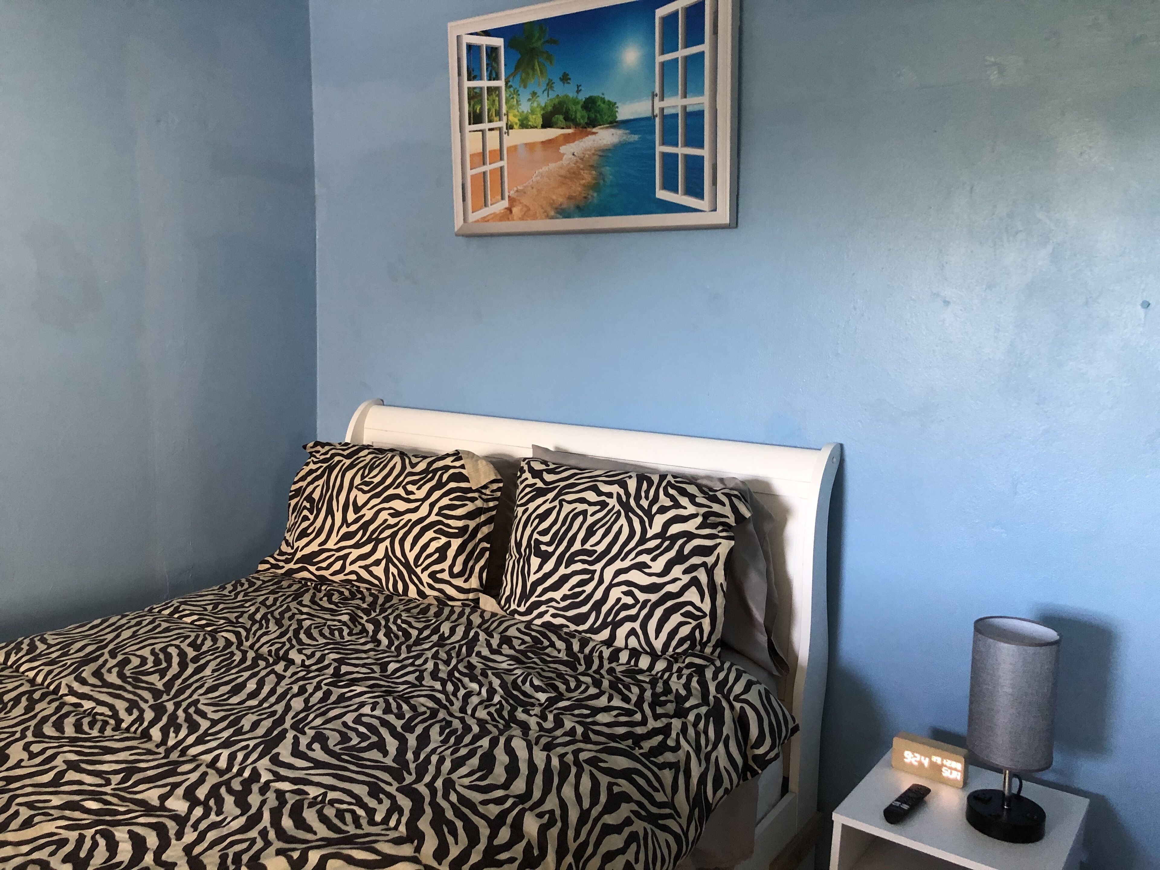 Un dormitorio con cama, almohadas de estampado de cebra, una mesita de noche con lámpara y un cuadro de una playa.