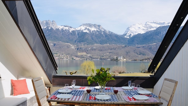 Ein Esstisch mit Geschirr, Gläsern und Blumen, mit Blick auf einen See und Berge.
