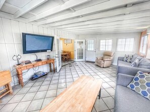 Ein geräumiger Wohnbereich mit einem Holztisch, einem grauen Sofa mit gemusterten Kissen, einem an der Wand montierten Fernseher und einem Essbereich, der durch eine offene Tür sichtbar ist.