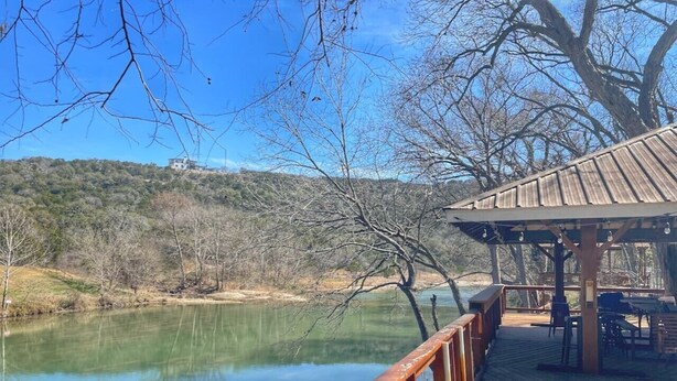Una terraza de madera con vistas a un lago tranquilo, con un pabellón y zona de estar al aire libre.