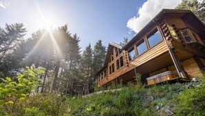 A wooden cabin with a balcony, surrounded by dense forest and sunlight streaming through the trees.
