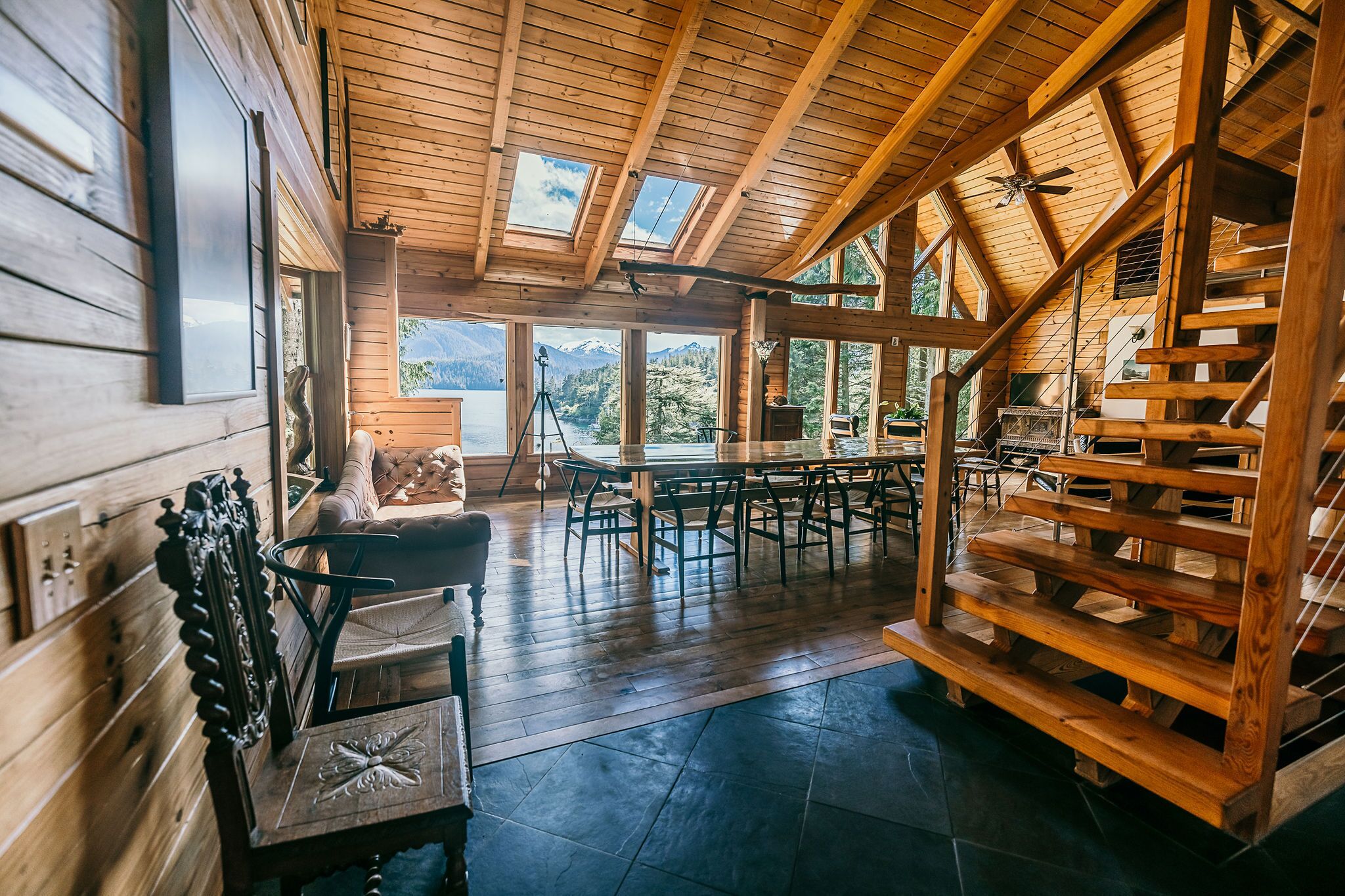 A spacious wooden interior with a high ceiling, large windows offering mountain views, a dining area, and a staircase.